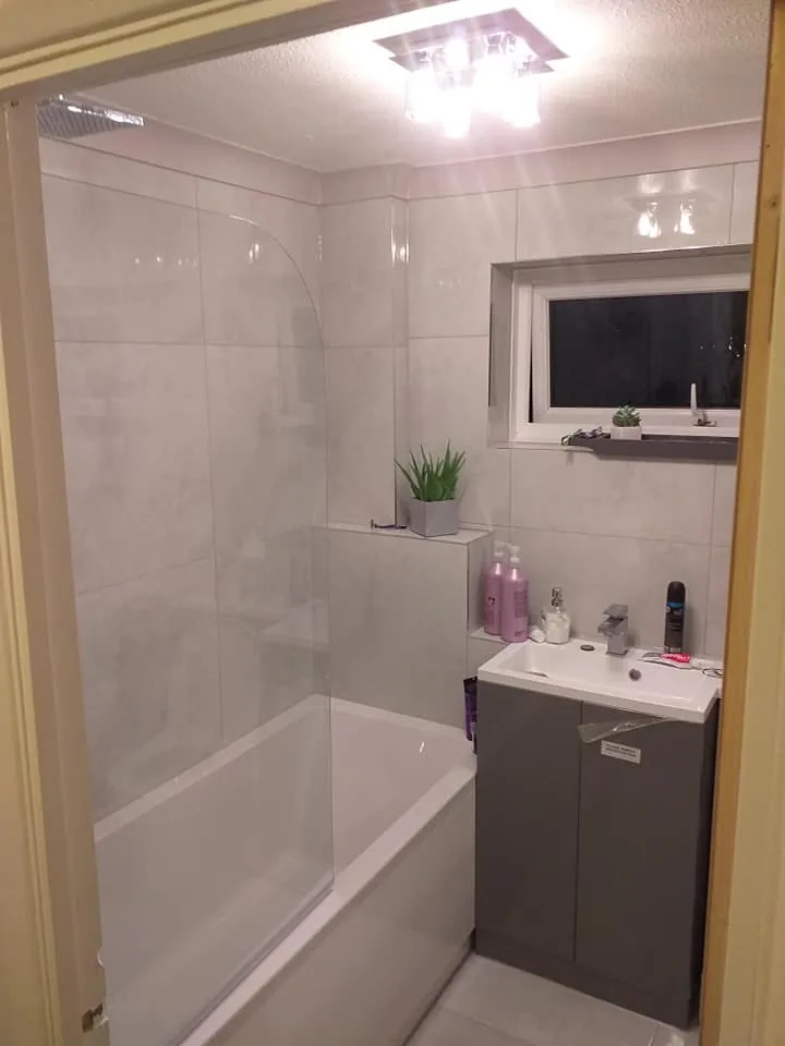 Modern bathroom with white tiled walls, a glass shower screen over a white bath, and a grey vanity unit with white basin.