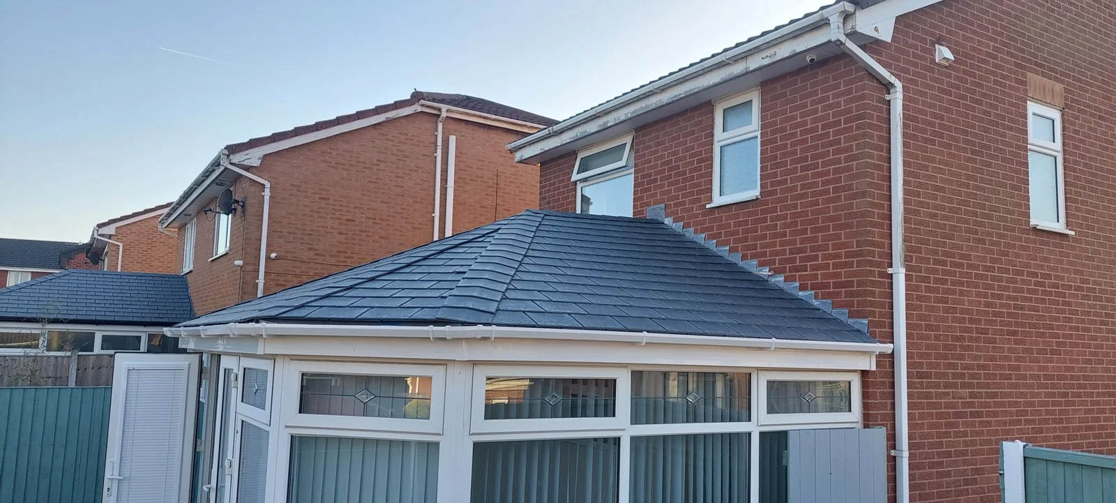 Newly tiled conservatory roof in slate-look tiles, fitted onto a red-brick semi-detached house under a clear sky.
