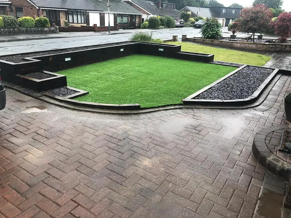 Front-garden landscaping: artificial lawn flanked by sleeper-edged borders with black slate, new block-paved driveway in the foreground.