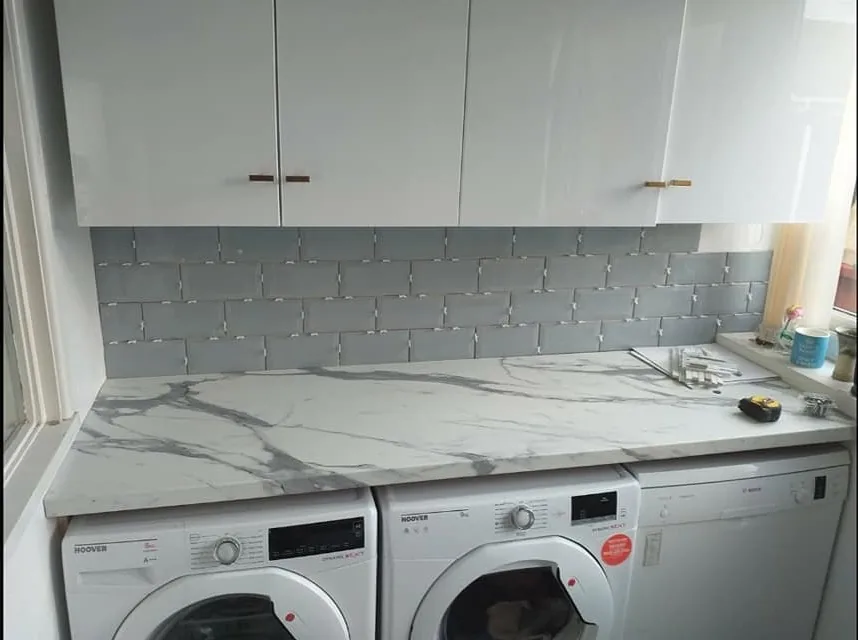 Utility kitchen with white gloss wall units, marble-effect worktop and grey metro-tile splashback above a washing machine and tumble dryer.
